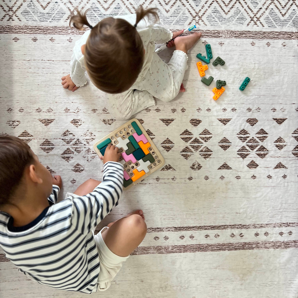 Two children playing with a wooden puzzle on a patterned rug.