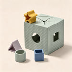 A pastel-colored geometric toy set on a warm beige background, featuring a light sage green cube with textured dots, a hole, and a window, alongside a yellow star, a blue square, and a lavender triangle.