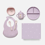 A coordinated baby feeding set arranged on a light gray background, including a lavender silicone bib with white star print, a matching plate, cup, spoon, fork, and square mat. The cup has a fox illustration, and the bowl has a deer illustration.