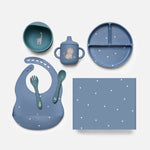 A coordinated children's dinnerware and feeding set arranged on a light gray background. The set includes a blue divided plate, a teal sippy cup with a lion illustration, a blue bib with white and gold speckles, a blue bowl with a giraffe illustration, matching utensils, and a square napkin or mat with white speckles.