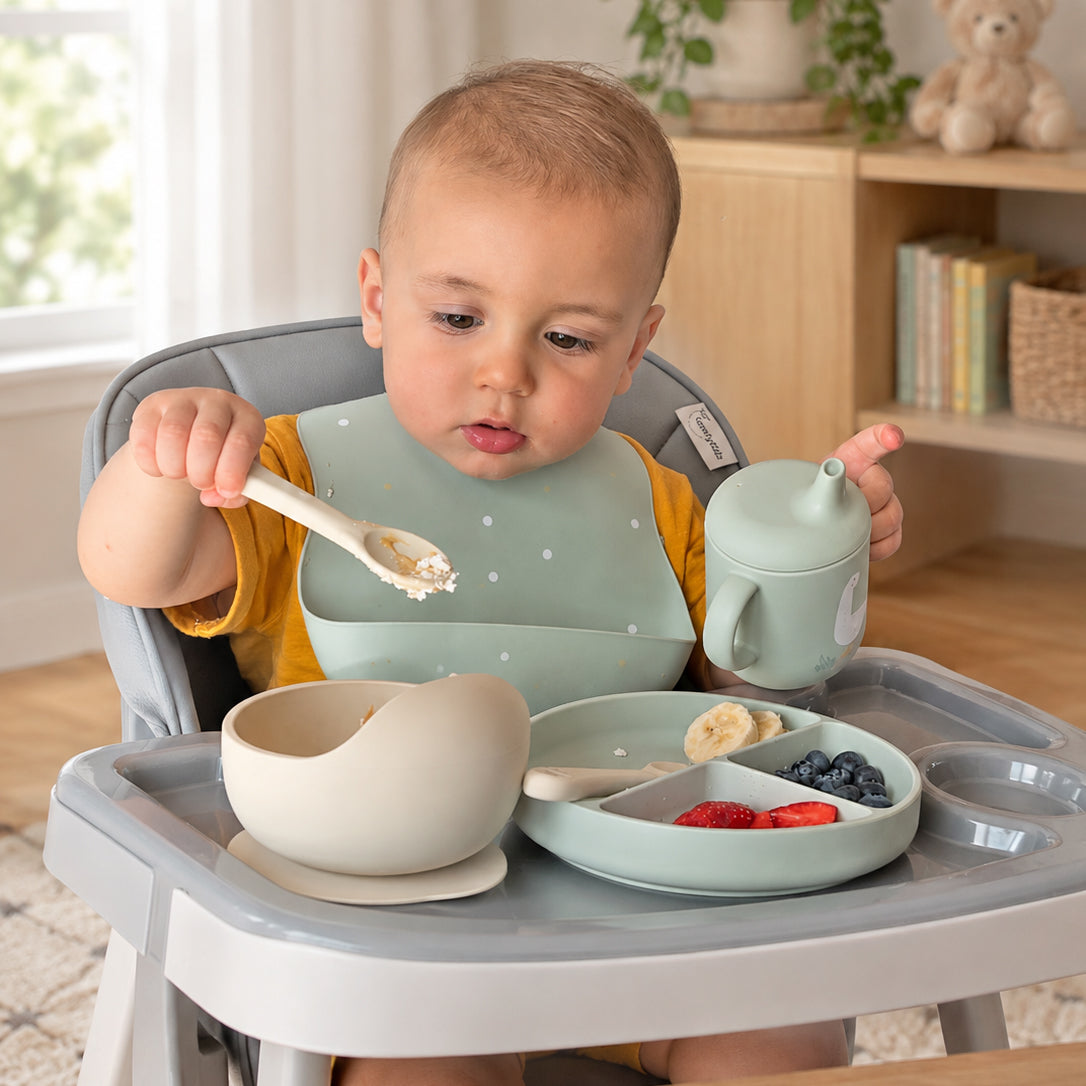 A baby sits in a high chair holding a spoon while eating with a matching light green bib, cup, bowl, and divided plate on a gray tray. The scene is set in a bright, modern nursery.