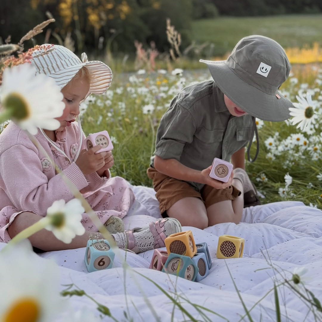 Two children sitting on a white quilted blanket in a field of white daisies, holding small wooden number blocks. The child on the left wears a pink outfit and striped sun hat, while the child on the right wears a gray-green shirt, brown shorts, and a gray bucket hat.