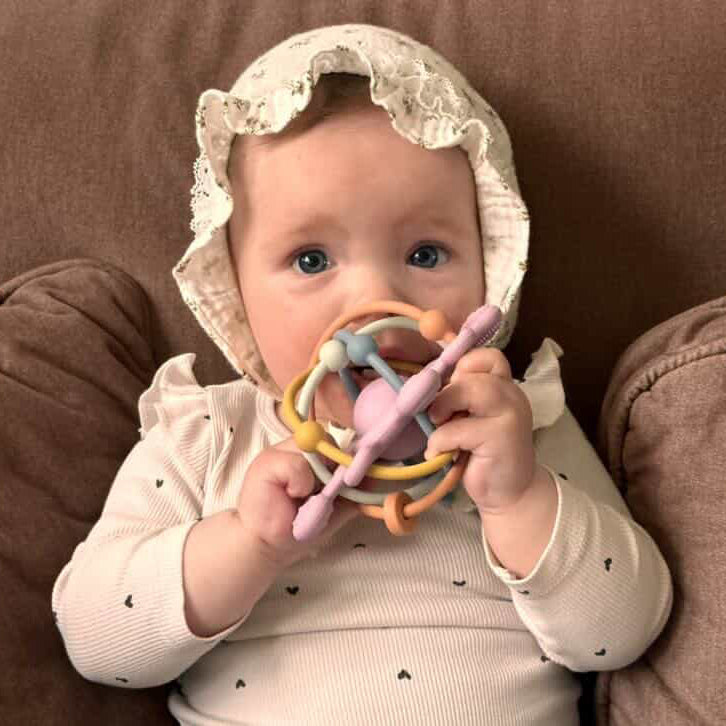 Baby wearing a lace bonnet holding a teething ring on a brown background