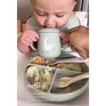 A baby is shown drinking from a sage green silicone cup with a goose illustration, held by an adult hand. The baby is seated at a table with a matching sage green divided meal tray and spoons, with a text overlay detailing product features.