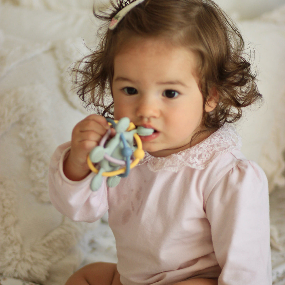 A toddler girl with curly brown hair wearing a light pink long-sleeve top with a lace collar, sitting on a white textured blanket and holding a pastel-colored fabric teether toy.