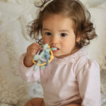 A toddler girl with curly brown hair wearing a light pink long-sleeve top with a lace collar, sitting on a white textured blanket and holding a pastel-colored fabric teether toy.
