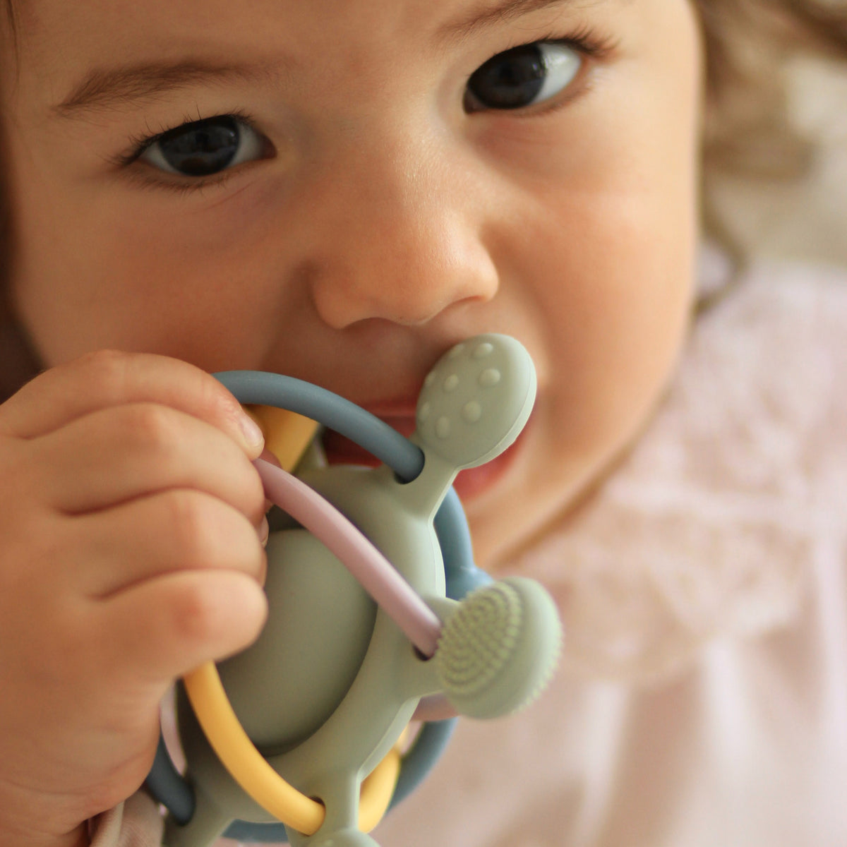 A close-up lifestyle shot of a baby's face and hand holding a pastel-colored silicone teether toy with a rattle and brush.