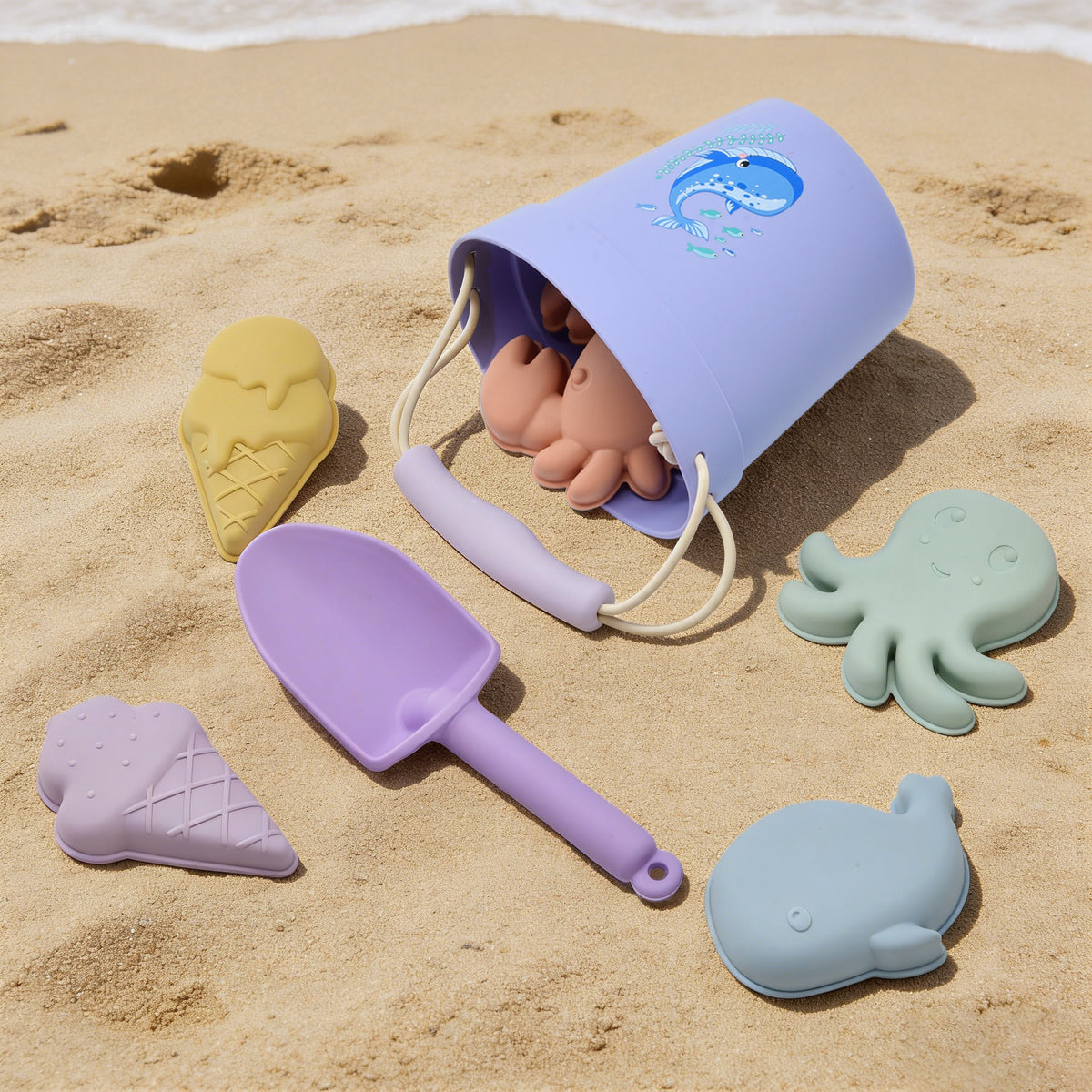Set of beach toys including a purple shovel, yellow mold, blue bucket, and green octopus toy on sand.  