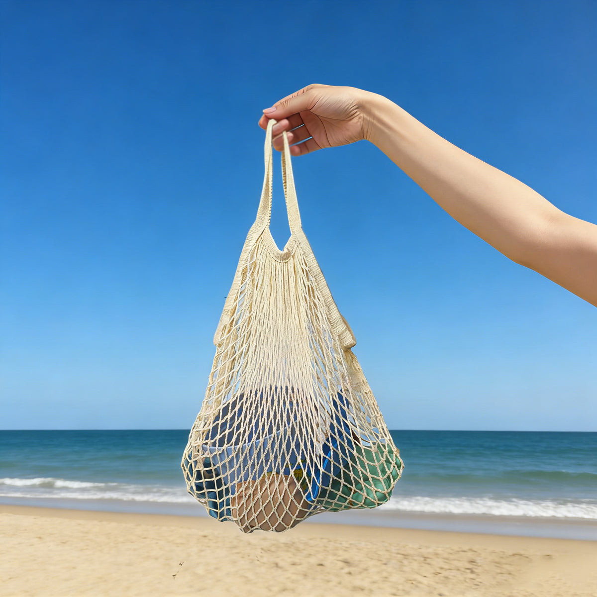 Hand holding a net bag with items on a beach against a clear blue sky