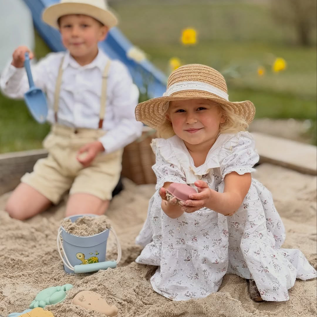 Two children playing in a sandbox with toys and sand, wearing hats.