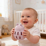 Baby holding a pink toy in a crib with toys and a teddy bear in the background 