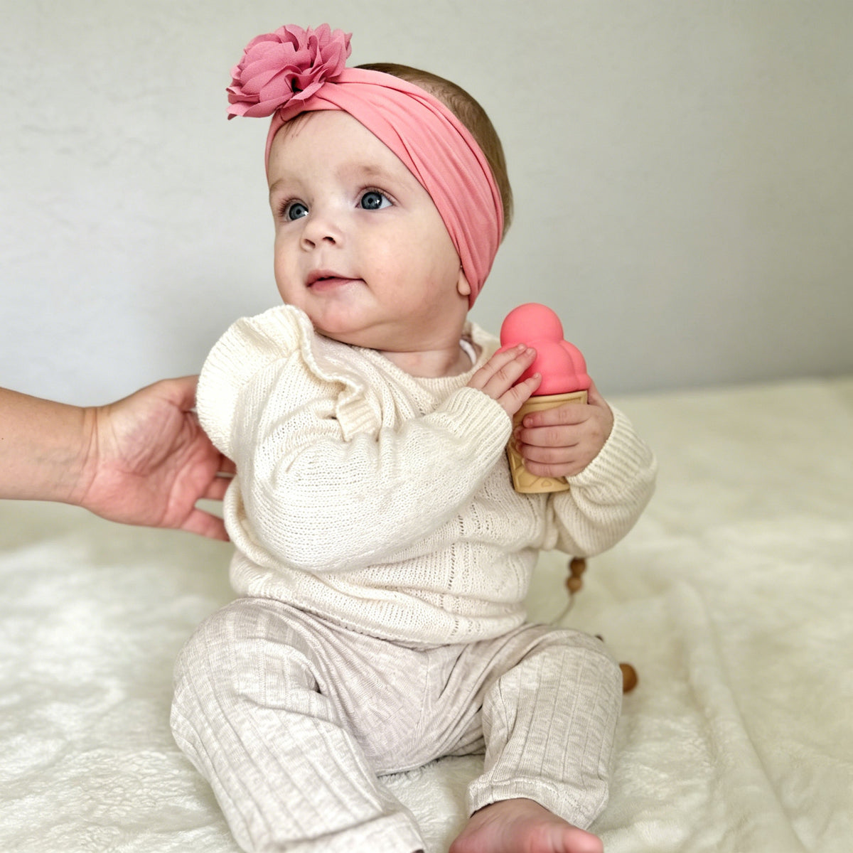 Baby wearing a pink headband and holding a pink toy, sitting on a white blanket.