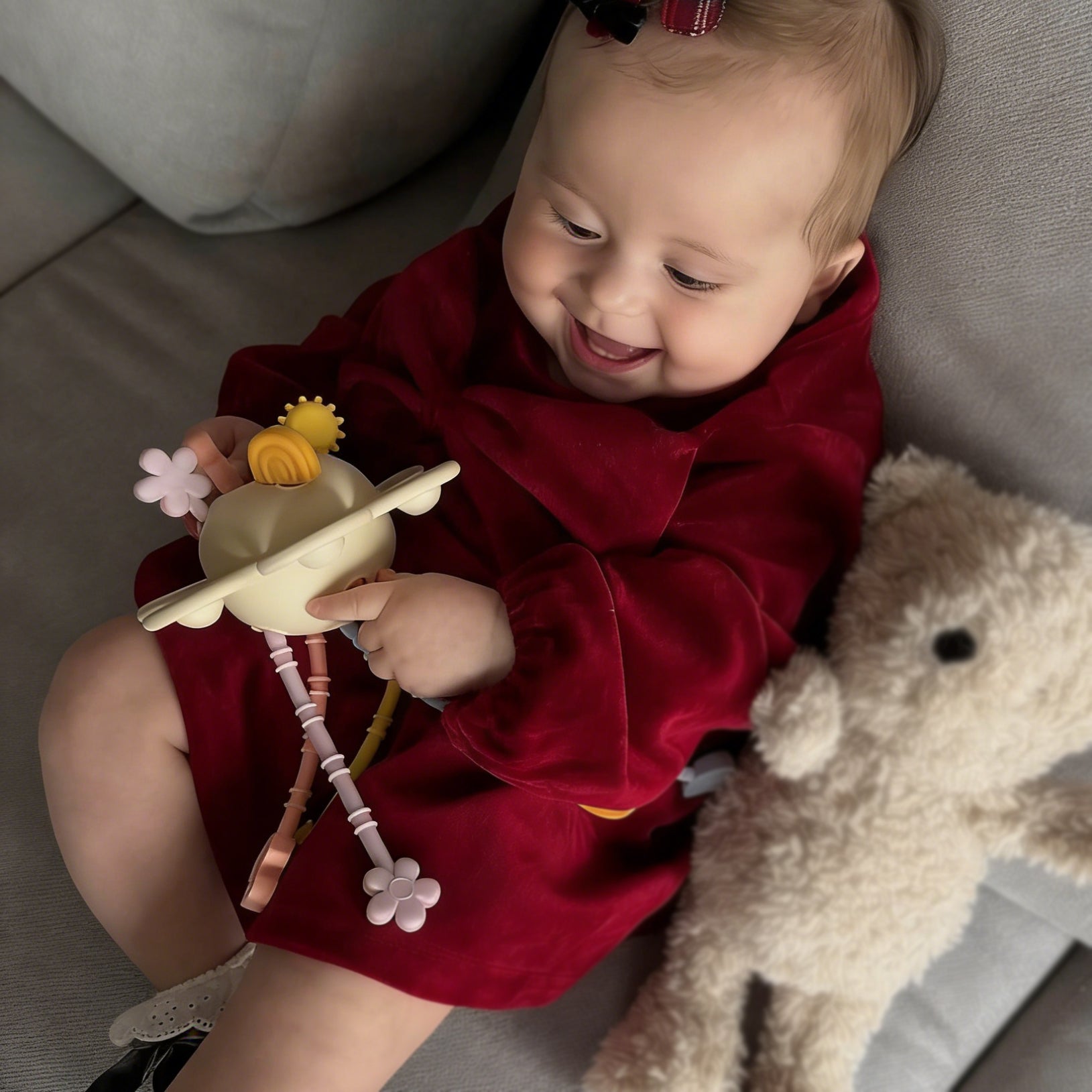 Baby in a red outfit holding a toy, sitting on a couch with a teddy bear beside.