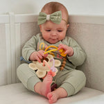 Baby sitting on a couch wearing a green outfit and headband, holding toys.