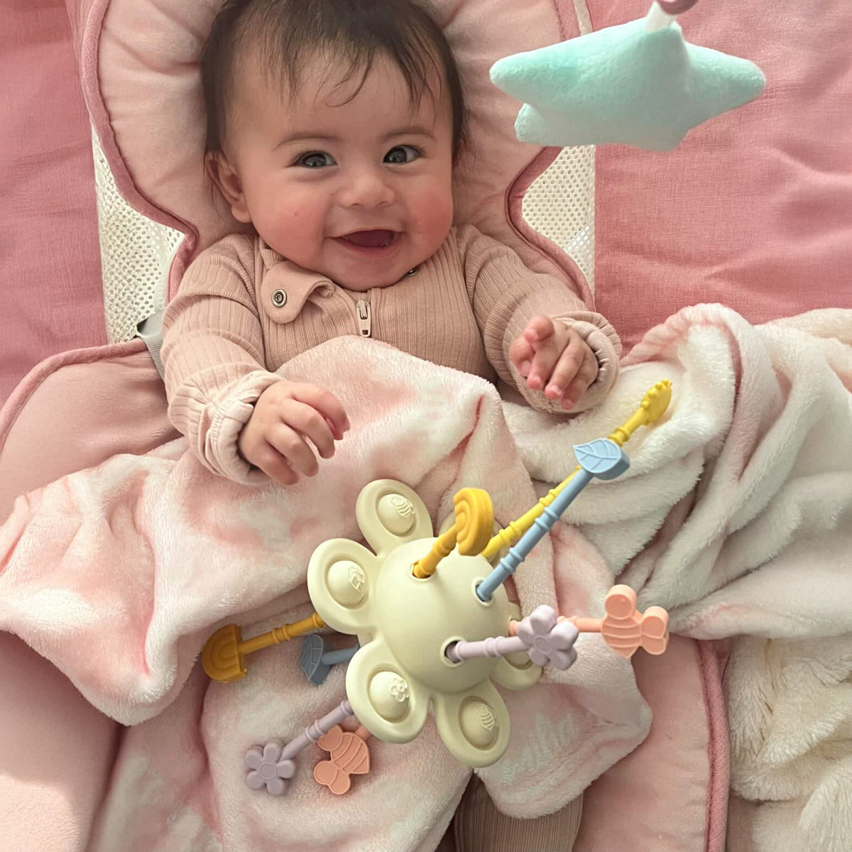 Baby lying on a pink blanket with a toy, smiling.
