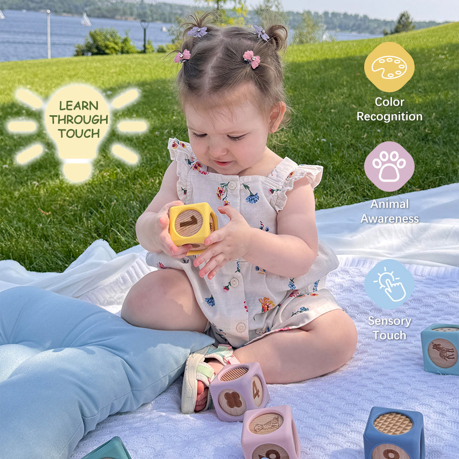 A toddler sits on a white blanket outdoors on a grassy lawn, holding a yellow block toy with a letter cutout. Other pastel block toys are scattered nearby, and text callouts on the right describe learning benefits such as color recognition, animal awareness, sensory touch, and learn through touch.