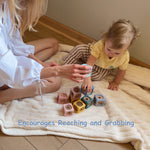A toddler in a yellow shirt and brown striped pants sits on a cream textured blanket on a wooden floor, reaching for colorful wooden blocks. An adult in a white blouse sits beside them, holding a matching block.