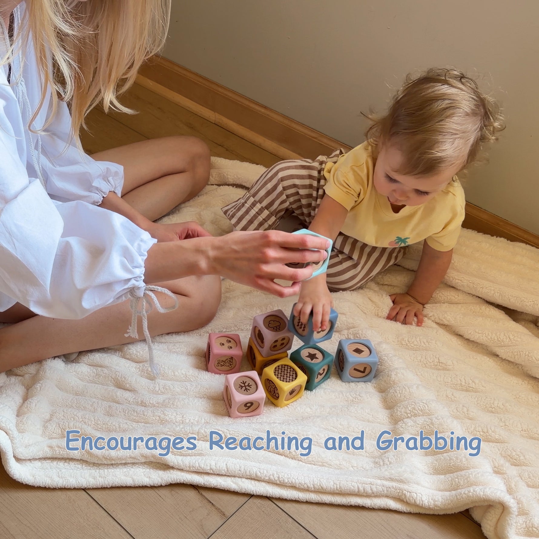 A toddler in a yellow shirt and brown striped pants sits on a cream textured blanket on a wooden floor, reaching for colorful wooden blocks. An adult in a white blouse sits beside them, holding a matching block.