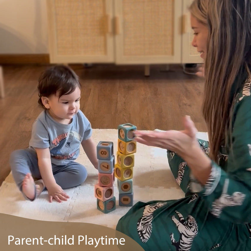 A toddler sits on a white play mat indoors, stacking colorful wooden number blocks while a woman in a green patterned robe watches and gestures. The scene is set in a home with wood flooring and woven cabinet doors in the background.