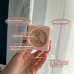 A hand holding a light pink silicone sand block with a natural wood lion engraving, set against a soft-focus background of curtains and a window. Text callouts highlight the product's features and dimensions.