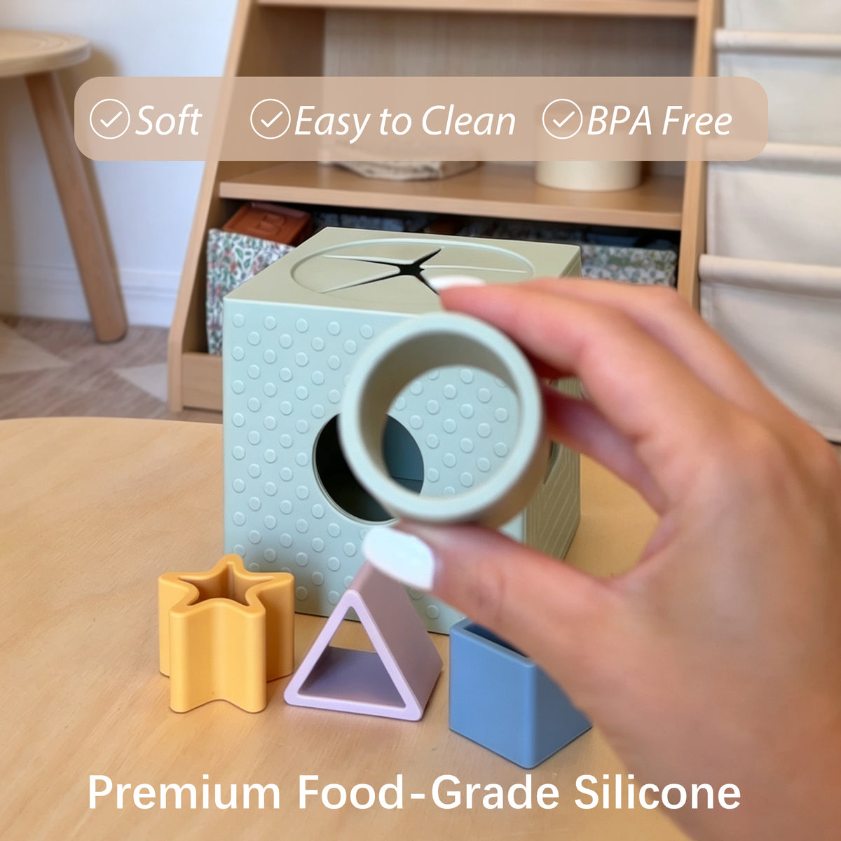 A hand holds a light sage green silicone ring mold over a wooden toy box with colorful geometric shapes, with text overlays highlighting soft, easy-to-clean, and BPA-free qualities.