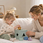 A toddler and a woman sit on a cream sofa playing with a pastel shape-sorting toy box. The child is inserting a yellow star shape into the box, which has black star, triangle, and square cutouts.