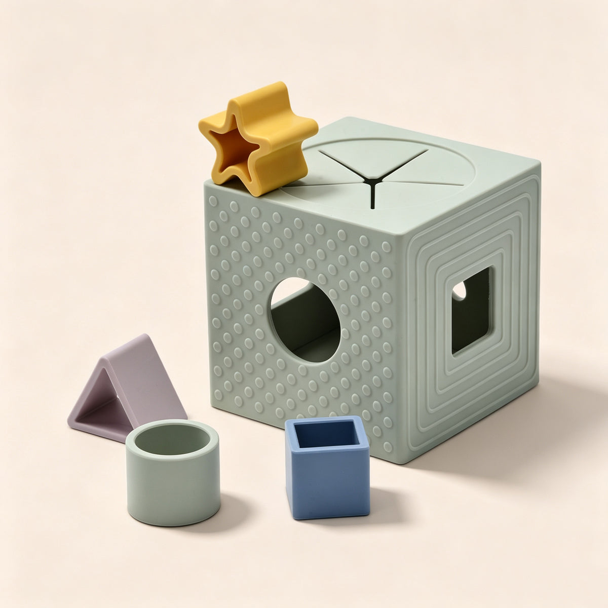 A pastel-colored geometric toy set on a warm beige background, featuring a light sage green cube with textured dots, a hole, and a window, alongside a yellow star, a blue square, and a lavender triangle.