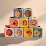 A stack of pastel wooden toy blocks arranged in a pyramid on a warm beige surface. The blocks are light blue, mint green, pink, yellow, and teal, each engraved with different wooden icons including paw prints, a triangle, the number 1, rain drops, a penguin, a heart, and a chevron pattern.