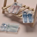 Set of children's cutlery with blue handles in clear plastic cases on a wooden stand.