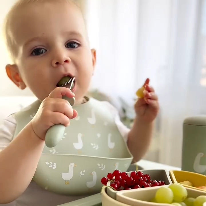 Baby eating with a spoon and holding a piece of fruit, wearing a bib with duck patterns 