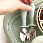 Children's spoon and fork set with sage handles held by a hand on a green tray 