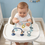 Baby sitting in a high chair with a colorful toy, in a room with light blue walls and white furniture.
