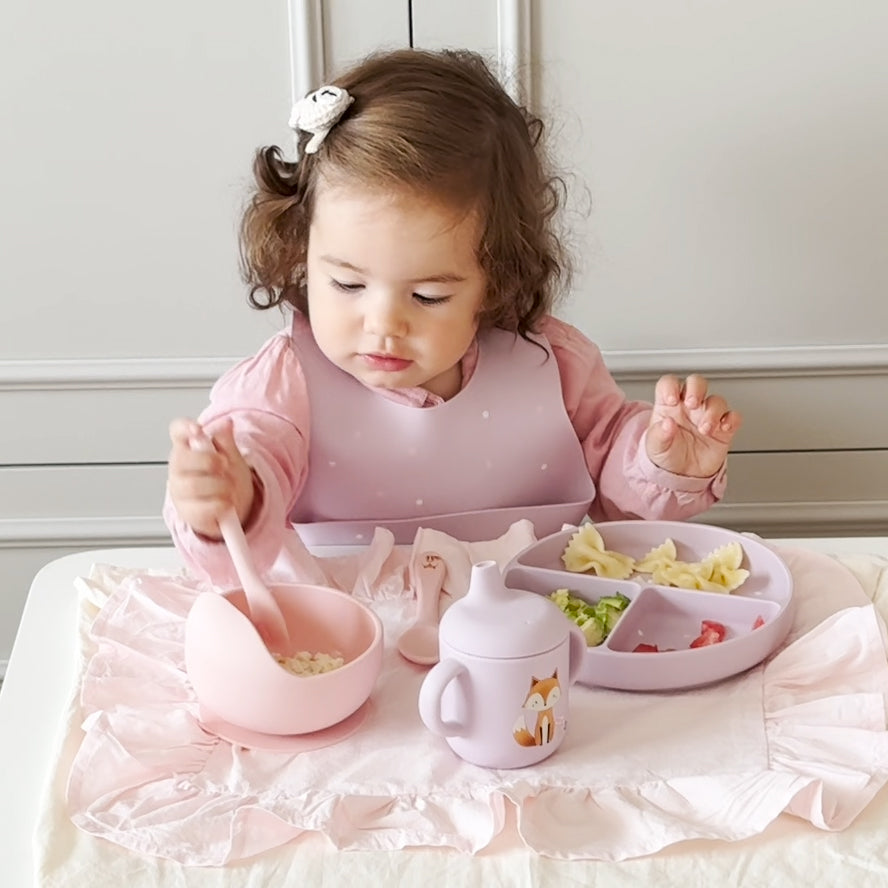 A toddler girl with curly brown hair wearing a pink long-sleeve top and a light pink bib sits at a table, eating from a matching pink bowl with a spoon, a divided plate with pasta and vegetables, and a small pink sippy cup with a fox illustration.