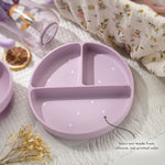 Purple children's plate with divided sections on a textured surface with flowers and a cup. 