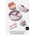 A 6-month-old silicone baby feeding set in soft pastel pink and lavender, including a divided plate, suction bowl, sippy cup, spoon, and fork, displayed on a light gray background with the text 'Everything they need for starting solids.'.