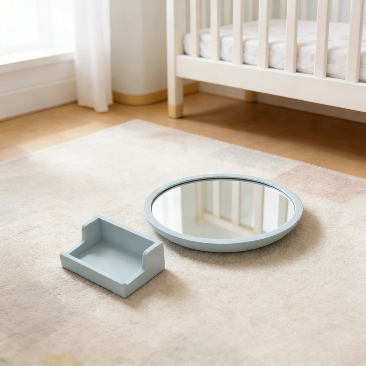 Round mirror and square tray on a beige rug in a nursery with a crib in the background. 