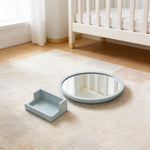 Round mirror and square tray on a beige rug in a nursery with a crib in the background. 
