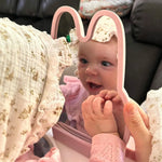 Baby looking at a pink mirror with bunny ears, wearing a pink outfit. 