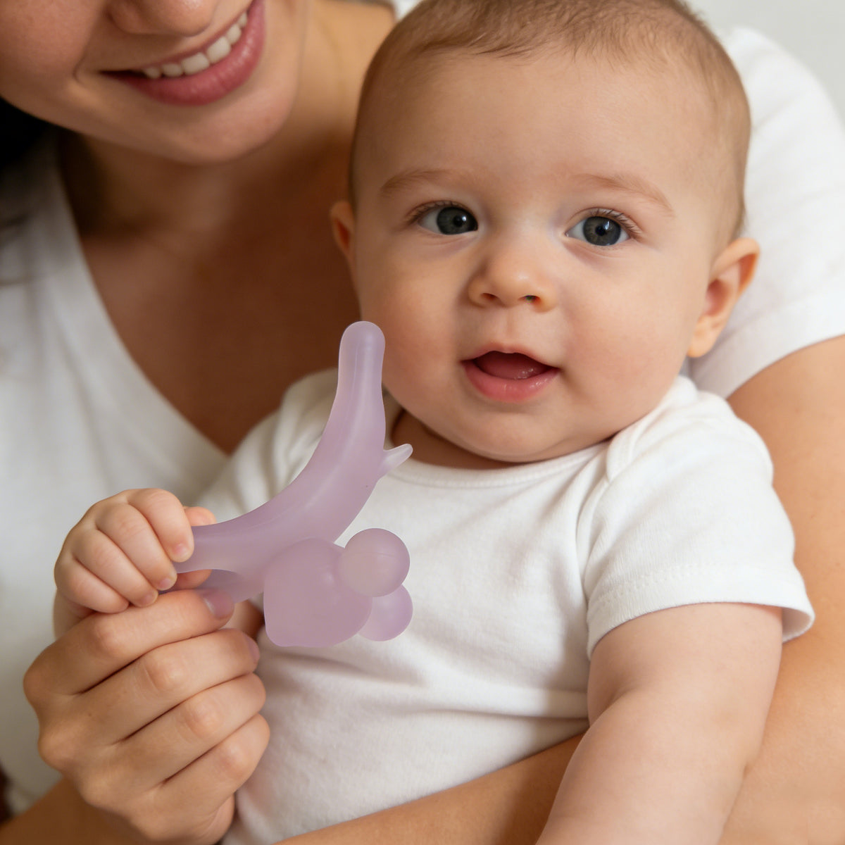 Baby holding a purple teething toy with a smiling woman in the background