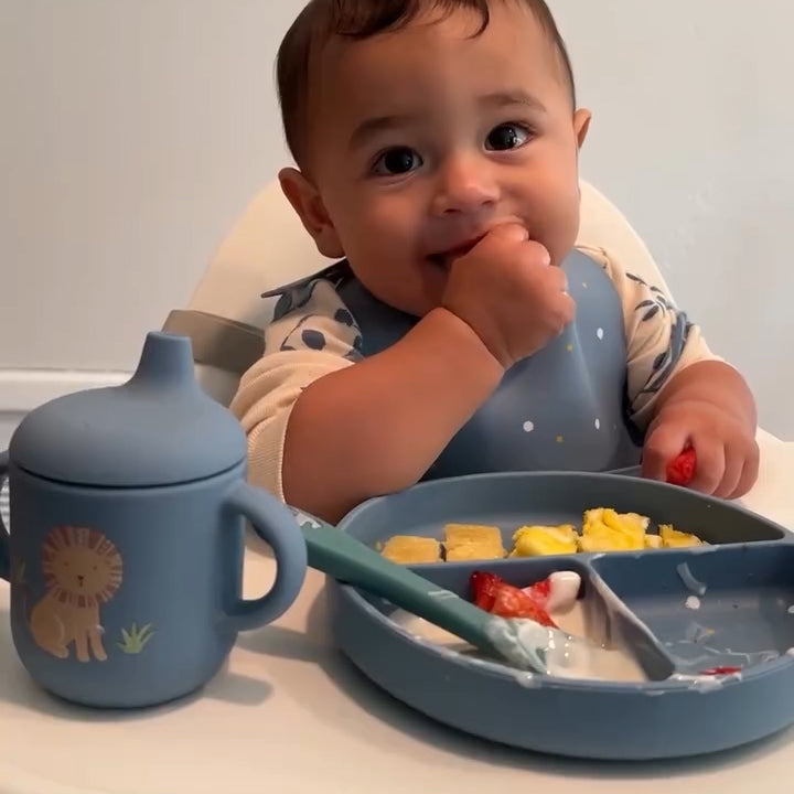 Baby sitting at a table with a blue sippy cup, plate, and bib.