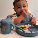 Baby sitting at a table with a blue sippy cup, plate, and bib.