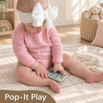 A baby wearing a pink long-sleeve bodysuit and a white bow headband sits on a cream woven rug, holding a Pop-It Play sensory toy. A brown banner at the bottom reads 'Pop-It Play'.