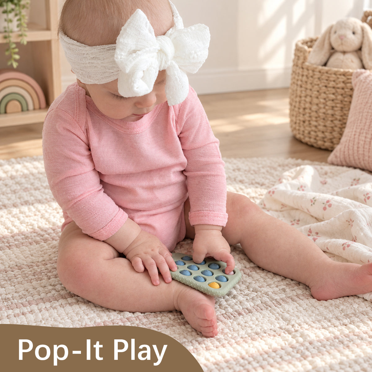 A baby wearing a pink long-sleeve bodysuit and a white bow headband sits on a cream woven rug, holding a Pop-It Play sensory toy. A brown banner at the bottom reads 'Pop-It Play'.
