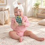 A baby wearing a pink outfit and a large white bow headband sits on a fluffy cream rug, holding and biting a mint green silicone toy with colorful circular buttons. The scene is set in a bright, sunlit nursery with wooden floors, a basket, and a plant in the background.