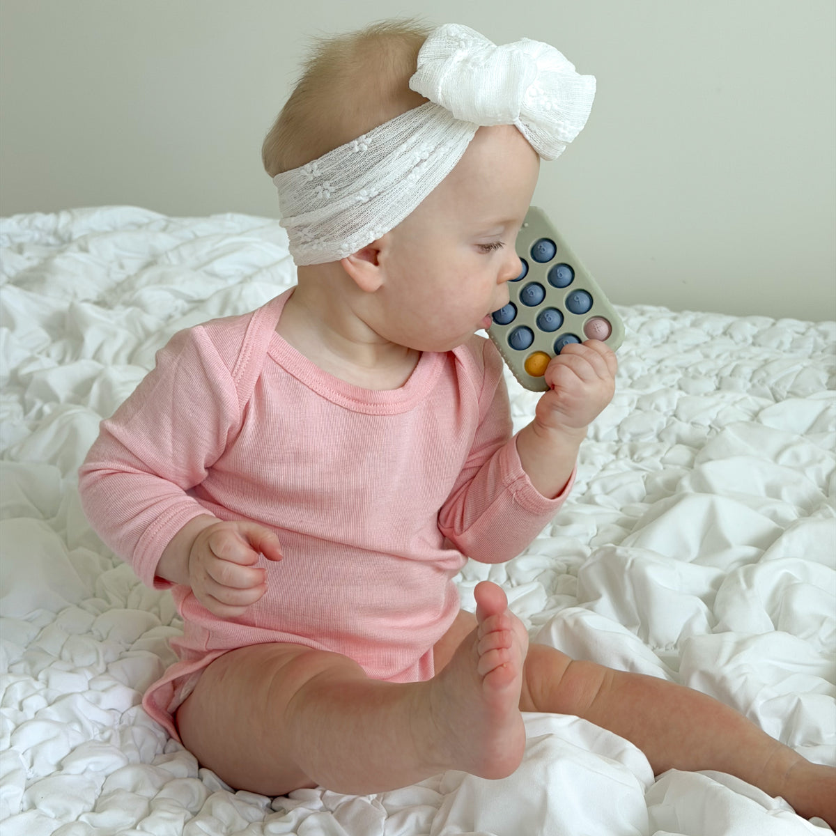 A baby girl wearing a pink long-sleeve bodysuit and a white lace headband with a bow sits on a white textured bed, holding a colorful pop-it toy to her mouth.