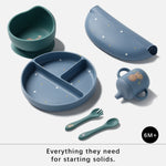 A 6-month+ baby feeding set with a heart-shaped bowl, divided plate, rolled lid, sippy cup, fork, and spoon in muted teal and dusty blue with small animal and star illustrations, shown on a light gray background with the text 'Everything they need for starting solids.'.
