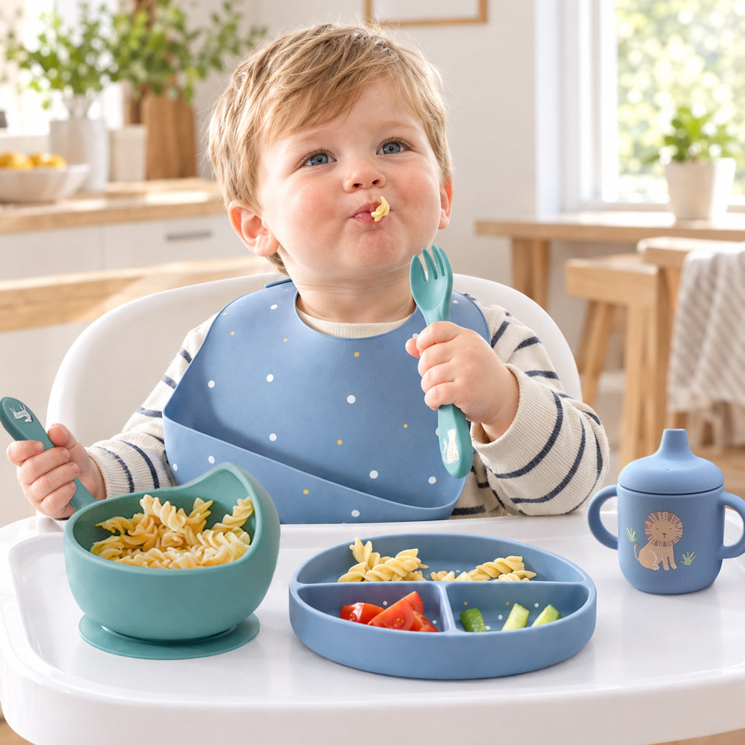 A toddler sits in a white high chair eating pasta with a blue silicone bib, matching blue spoon and fork, a blue bowl, and a divided plate with a blue sippy cup in a bright kitchen setting.