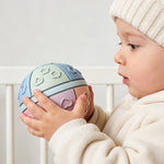Child holding a colorful ball with shapes, wearing a cream hat and sweater.