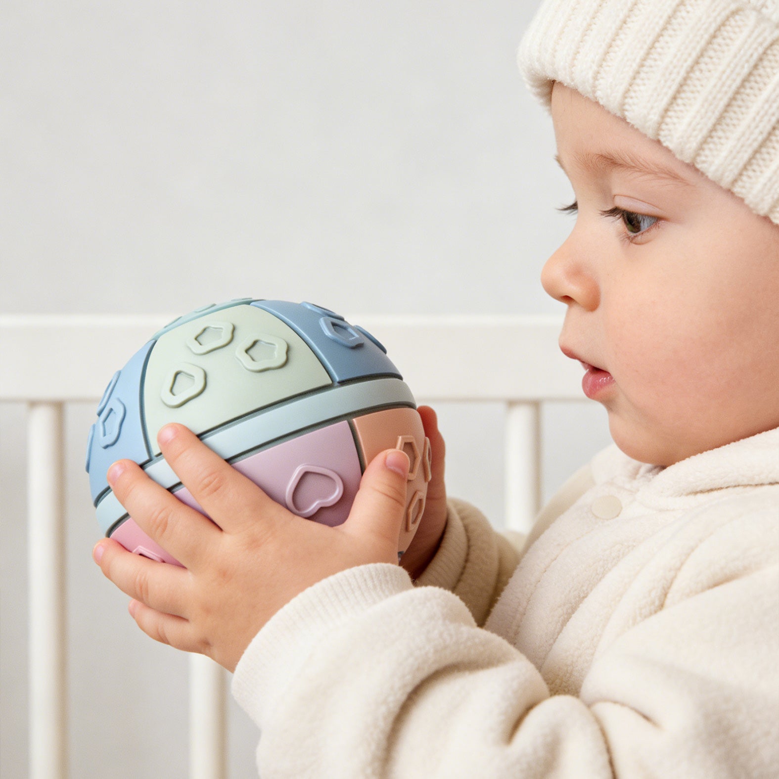 Child holding a colorful ball with shapes, wearing a cream hat and sweater.