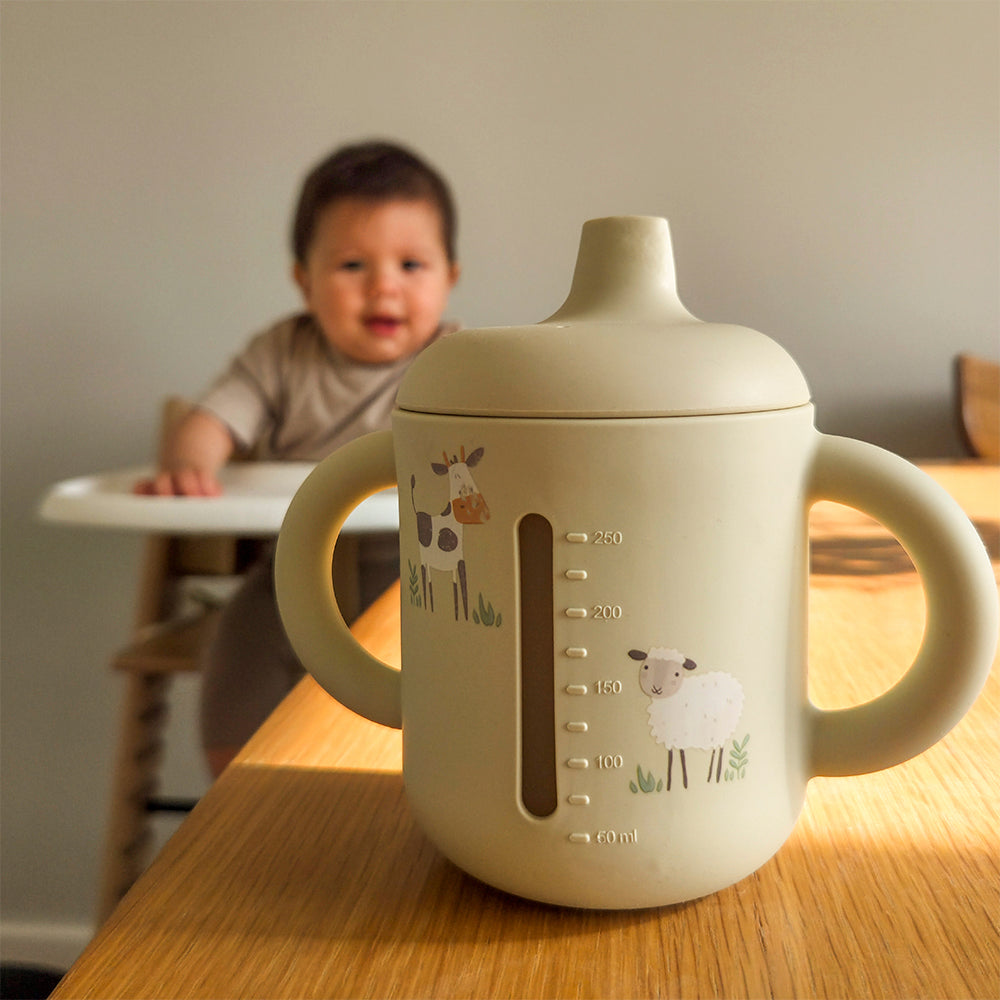 Children's sippy cup with animal illustrations on a wooden table, with a child in the background. 